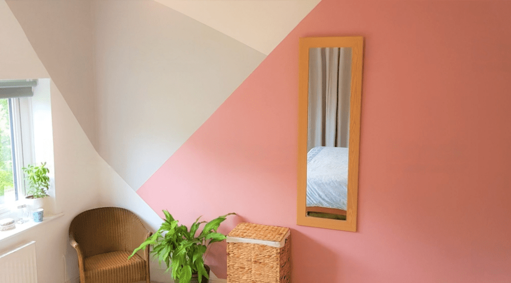 A room wall painted with pink  color having mirror on it and having the chair and plant in the room